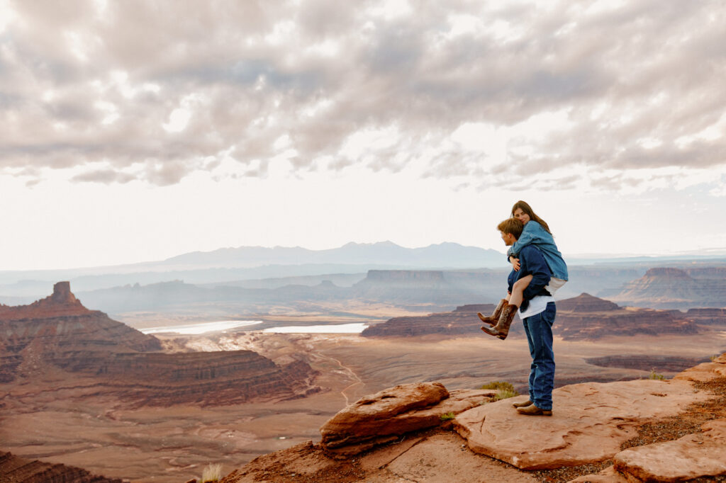 A bride is riding piggy back on the grooms back. They are both taking in the view of Dead Horse State Park during the sunrise portion of their elopement in Moab