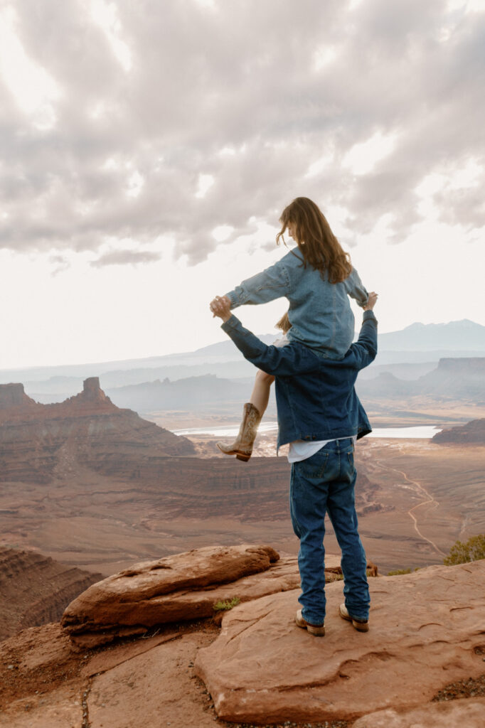 A bride sits on the shoulders of her groom, and holds his hands. They are both taking in the view of Dead Horse State Park during the sunrise portion of their elopement in Moab