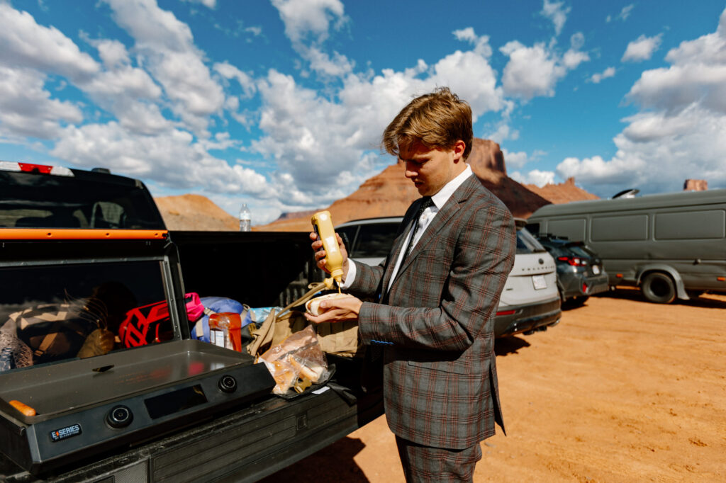 A groom puts mustard on a hot dog while he gets ready for his elopement.