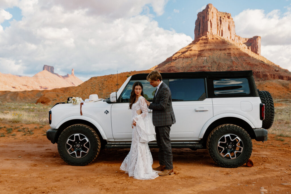 A groom helps his bride button her dress on their elopement day. They are standing in front of their Ford Bronco at Ida Gulch in Moab