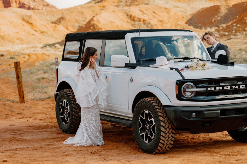 A bride and groom use the reflections of their Ford Bronco as mirrors to help them get ready for their elopement day.