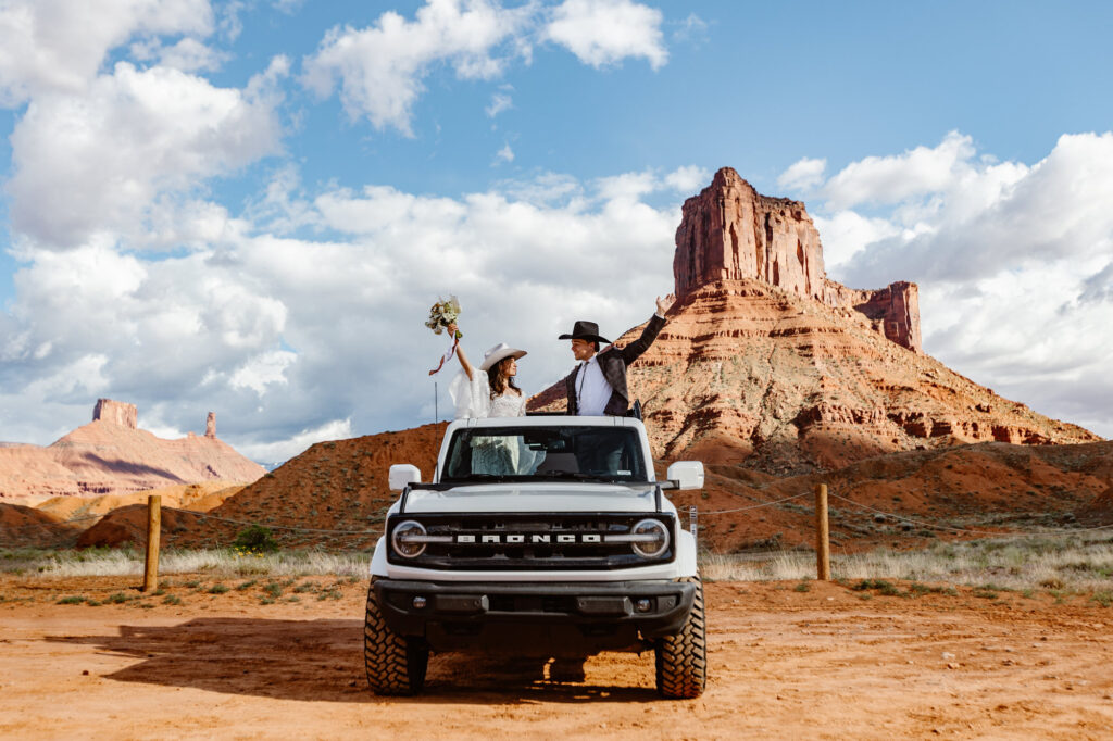 An eloping couple stands out of the sunroof of their Ford Bronco with their arms up. They are right by Ida Gulch Trailhead in Moab getting ready for their ceremony. They are both wearing cowboy hats.