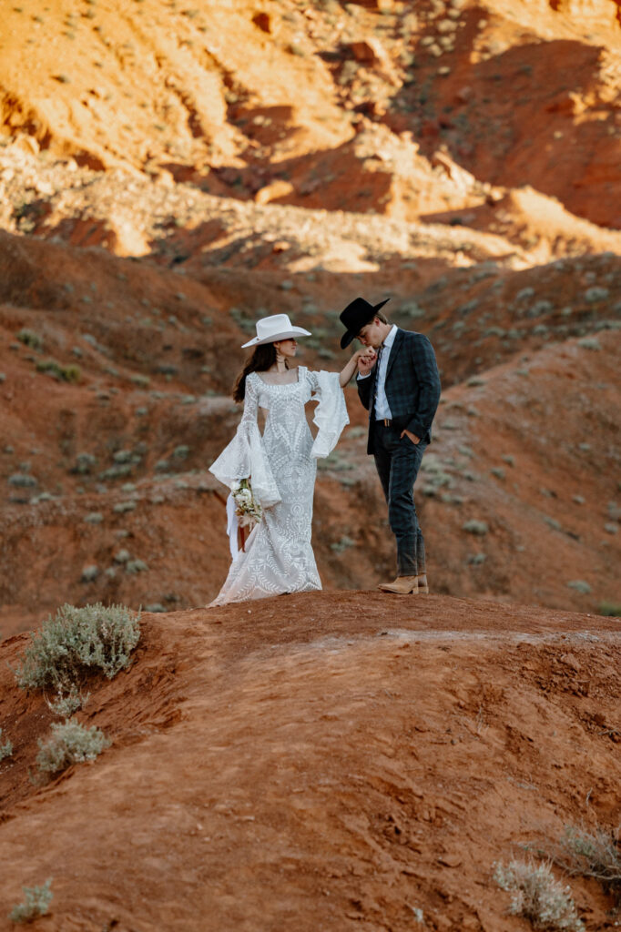 A bride and groom wearing cowboy hats stand in from of some red rocks in Moab. The groom is gently kissing the brides hand