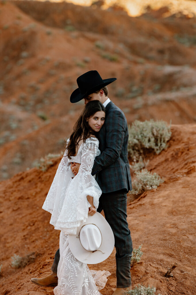 A photo of a bride and groom on their elopement day in Moab. He is wearing a cowboy hat, and she is holding a cowboy hat, He is nuzzled into her head, and she is looking at the camera