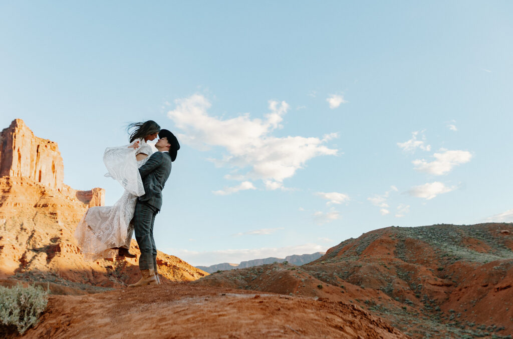 A groom picks up his bride during their elopement in In Moab. They are standing in front of some huge red rocks.