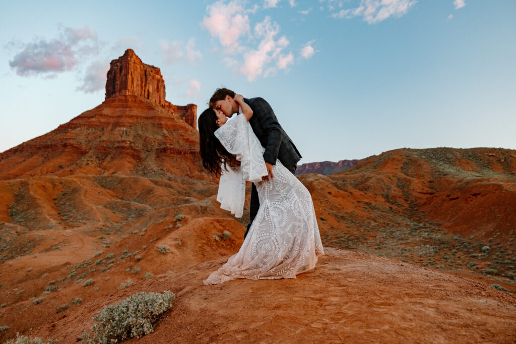 A bride and groom kiss at the end of the ceremony of their elopement. They are standing in front of some massive red rocks in Moab,