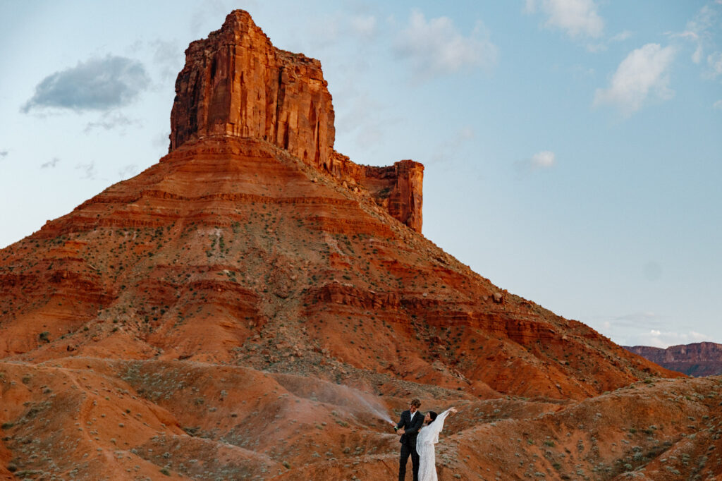 A bride cheers at her groom pops and sprays champagne celebrating their elopement. They are standing in front of some epic red rocks at Ida Gulch