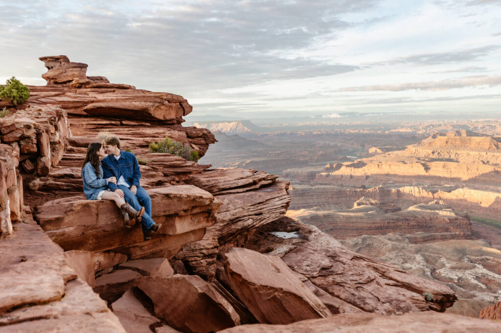 A couple sits on a red rock ledge at Dead Horse Point in Moab. The couple is wearing denim for their sunrise elopement session.