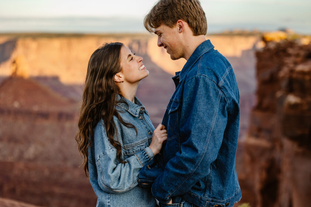 A couple stands facing each other and smiling at Dead Horse Point in Moab, They are both wearing denim jackets