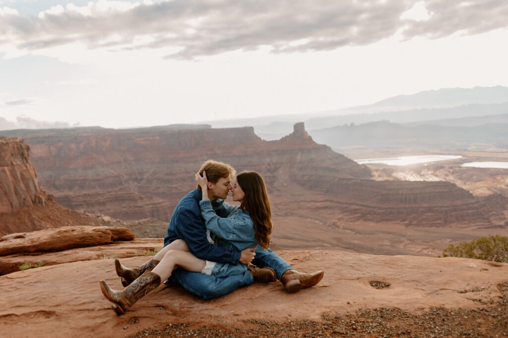 A couple is sitting on a red rock at Dead Horse Point in Moab. The bride has her legs over the grooms, and her hands ar wrapped around his neck. They are both wearing denim