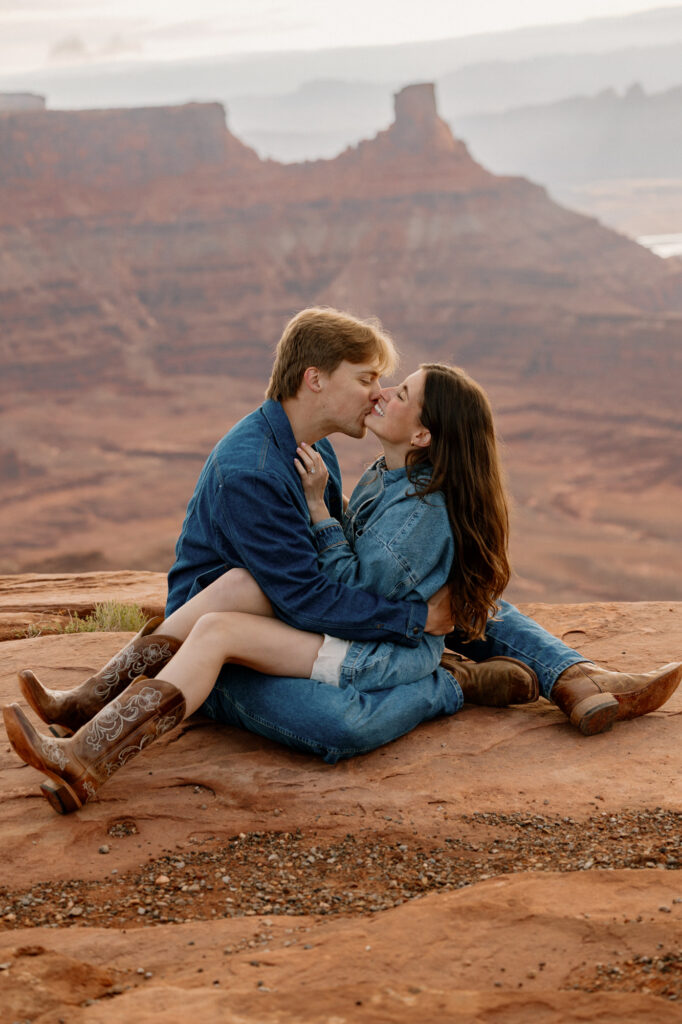 A couple kissing at the sunrise part of their elopement. The birds has her legs over the grooms, and they are kissing. Dead Horse State Park is in the background.