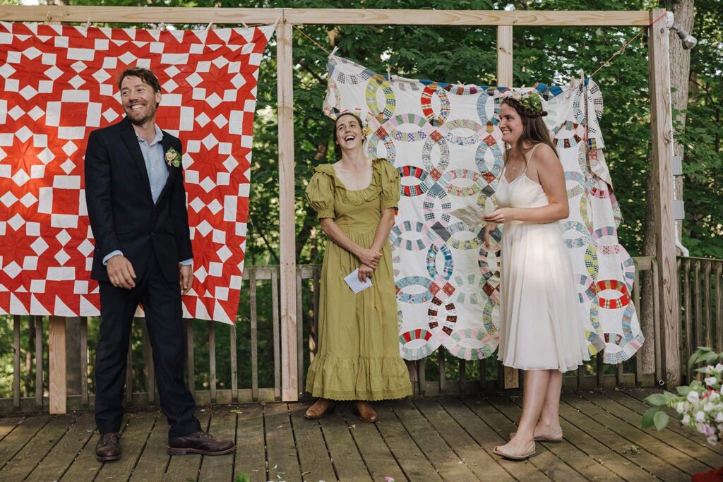 A bride and groom stand in front of two quilts made by the bride. The quilts are the background for their wedding ceremony, The couples is laughing as they read their vows. how to legally get married in pennsylvania