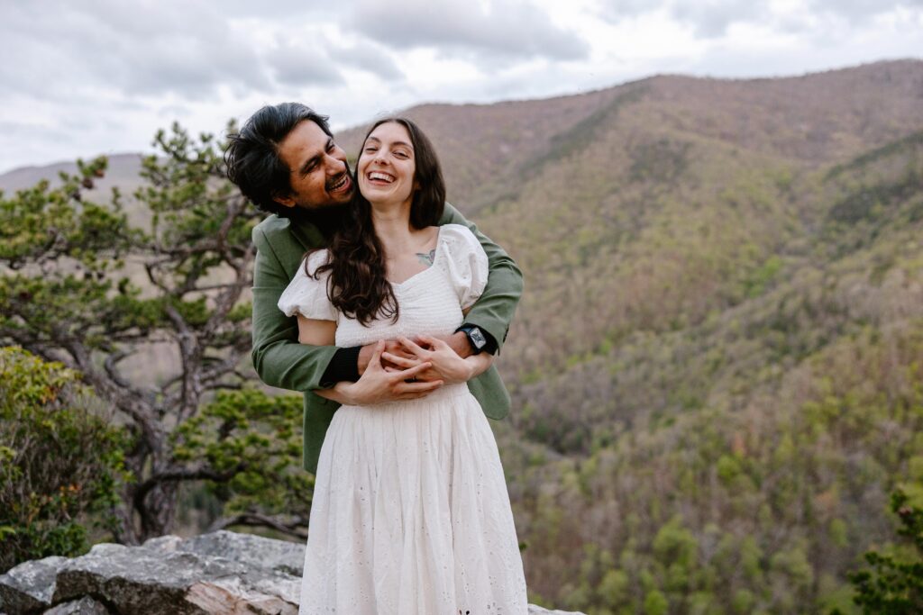 A groom has his arms around the bride, and they are both laughing. You can see the Virginia Mountains in the background