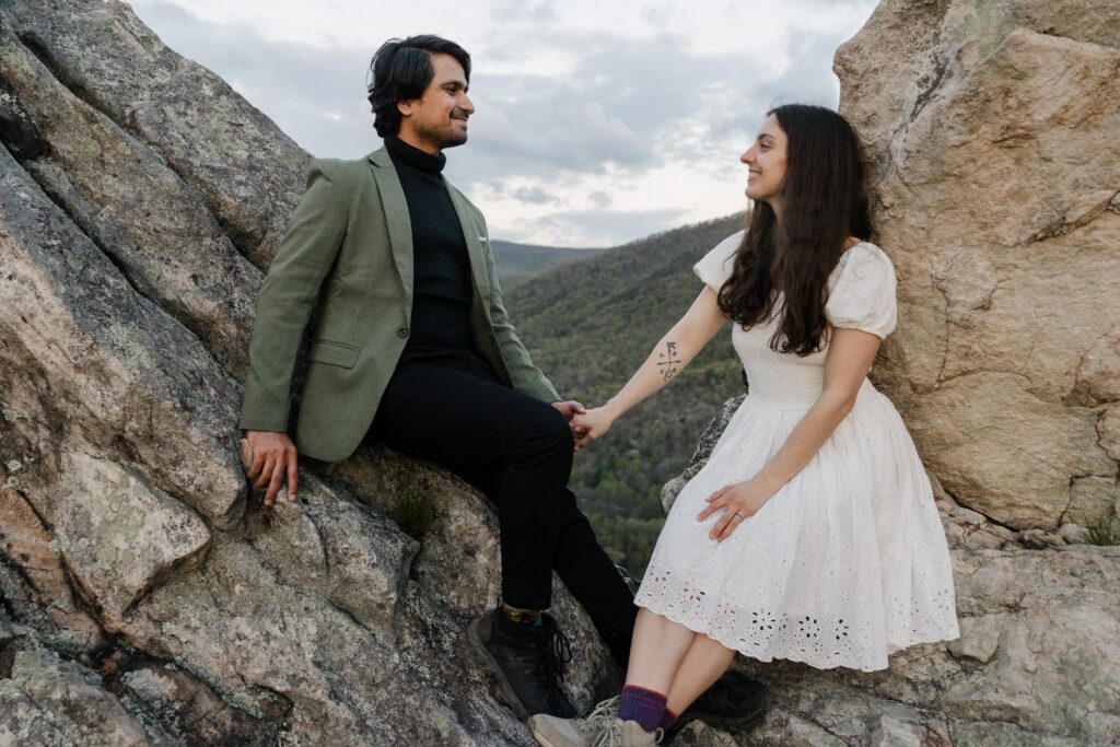 A bride and groom sitting on a giant rock formation on their sunrise elopement. They are holding hands, and smiling at one another. n