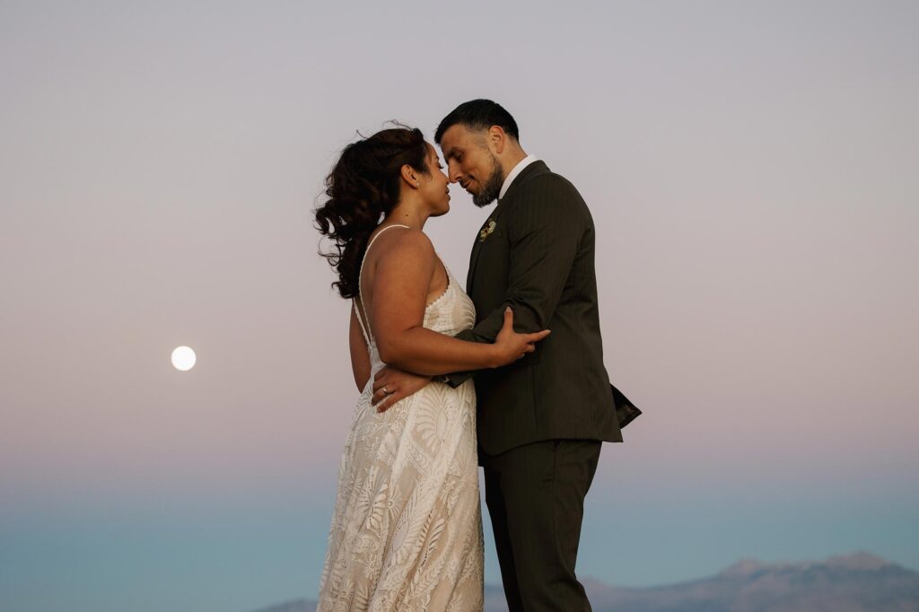 A bride and groom stand facing each other just about to kiss in Moab as the sunsets and the moon rises during their elopement.