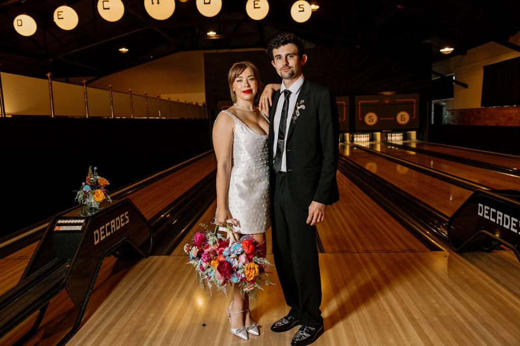 A bride and groom stand in the lane of a bowling alley at a vintage arcade during their elopement reception. The. bride is carrying a purse bouquet that is full of bold and colorful blooms