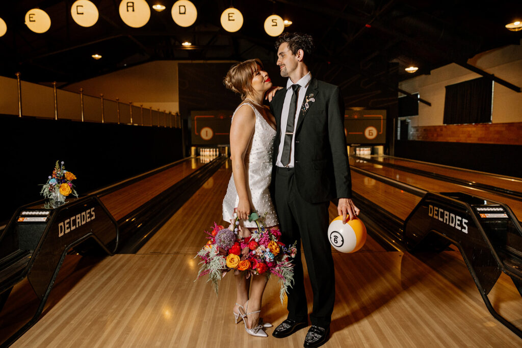 A birde and groom stand in the middle of a bowling alley during their non-traditional reception at a vintage arcade. The bride is wearing a shote dress with pearls, and holding a bouquet that is a purse, and the groom is holding a bowling ball