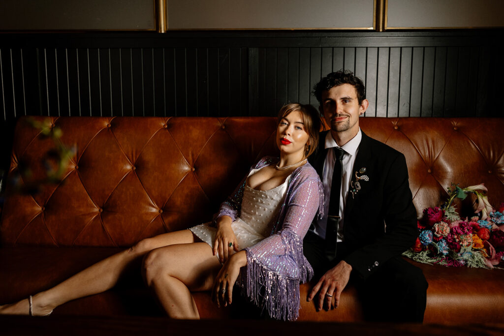 A bride and groom lounge on a couch during their reception. the bride is wearing a shirt dress and a purple sparkly finged shaw