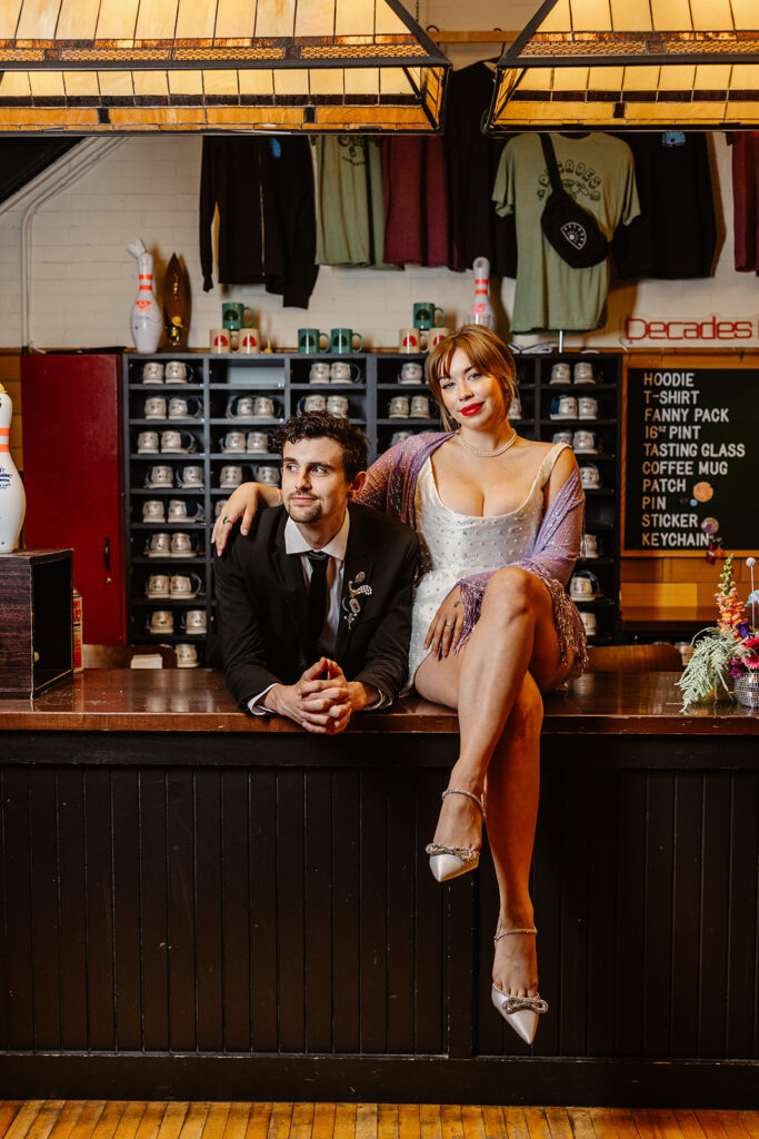 A bride and groom sit at the counter of the bowling alley section of the vintage arcade where they are holding their elopement reception. The bride is sitting on the counter with her arm around the groom. they are sitting in front of a rack of bowling shoes.