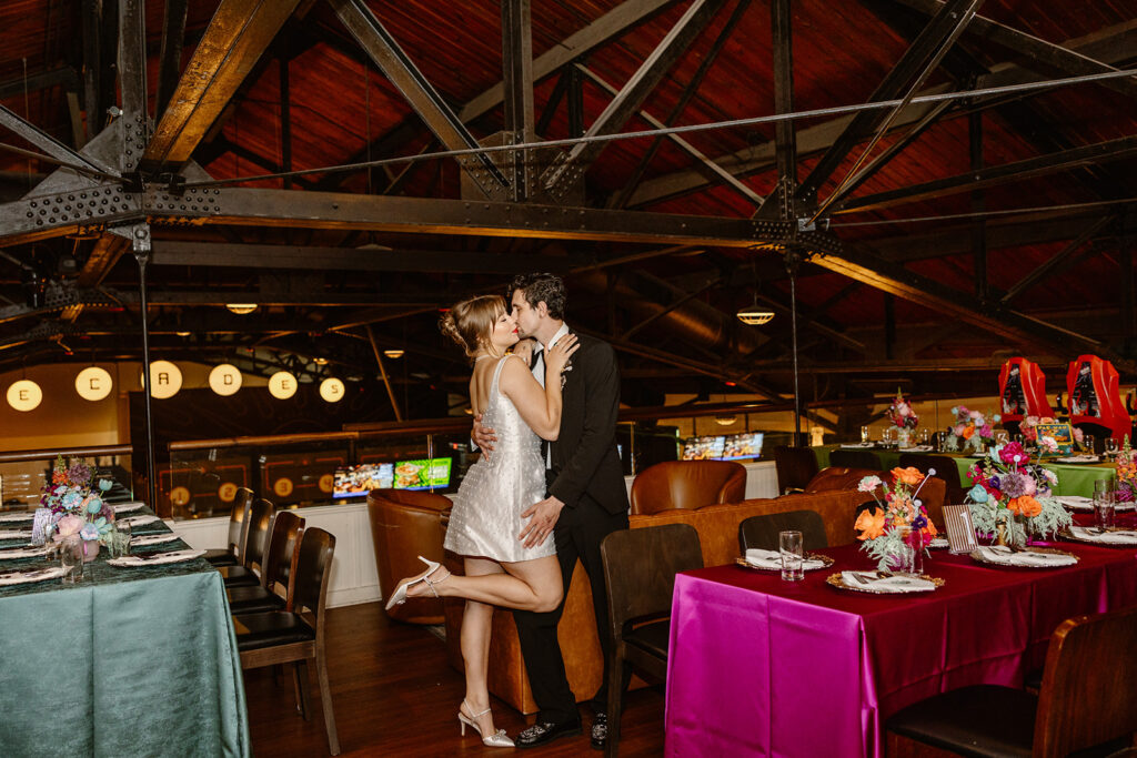 A bride and groom kiss while standing in front of the tables at their non tradtional wedding reception.