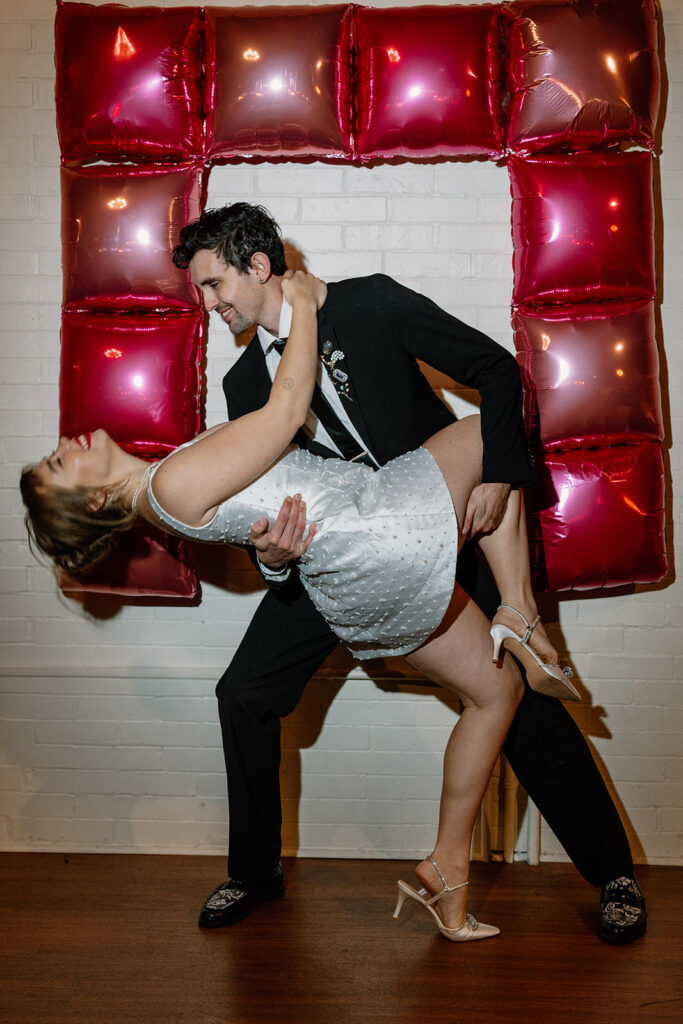 A groom dips his bride in front of some square shiny ballons on their wedding day