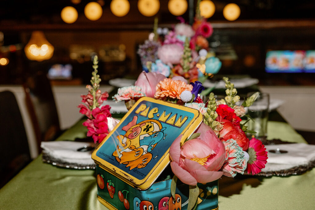 A photo of a bold and unique table scape at a fun vintage arcade reception. Featured is a vintage Pac-Man lunchbox with colorful flowers inside of it. 