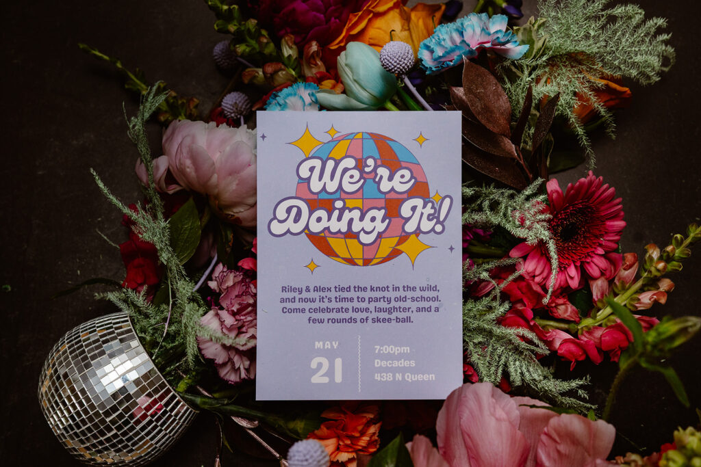 Photo of an invitation for a non traditional wedding reception at a vintage  arcade in Lancaster Pa. The photo has an invite with a colorful disco ball, and there aere flowers behind it