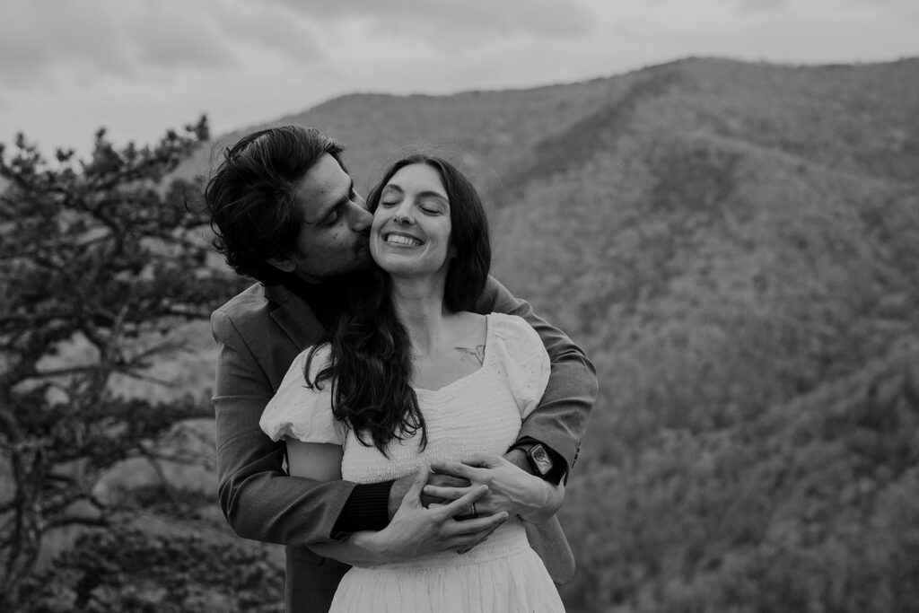 A groom snuggles behind his bride and wraps his arms around her in front of mountains on their wedding day.