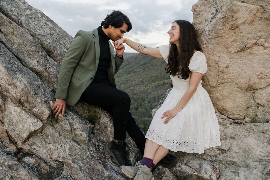 A groom kisses the hand of his bride while they sit on rocks at an overlook at Buzzard Rock in Virginia