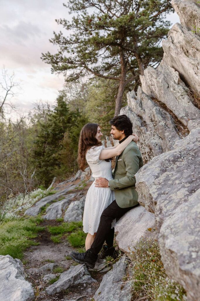 A groom sits on a rock, as his bride leans into him with her arms around him. They are looking at one another lovingly