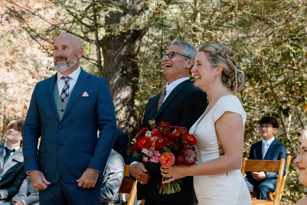 A bride and her stepfather meet up with the groom in the aisle during their wedding ceremony. All three of them are laughing and smiling.