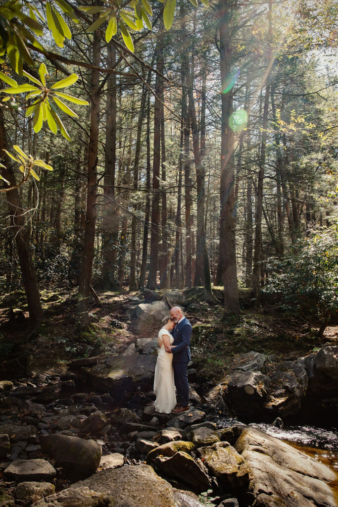 A bride and groom hug while standing in the middle of the forest with the sun shining through on their wedding day