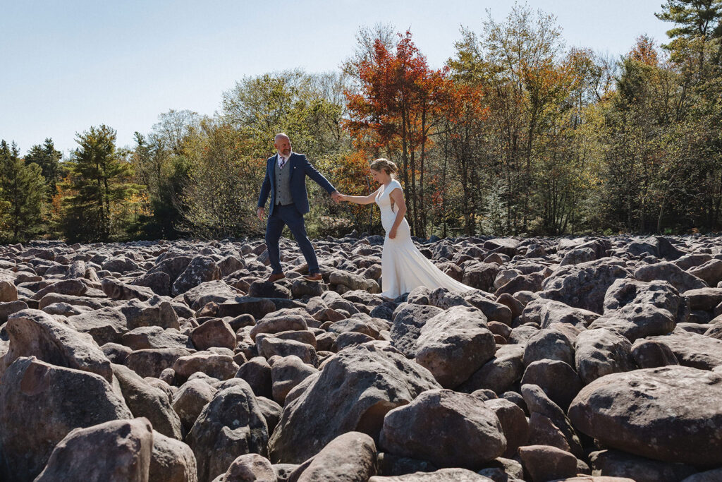A groom holds his wife's hand as he helps her walk across a boulder field on their wedding day.