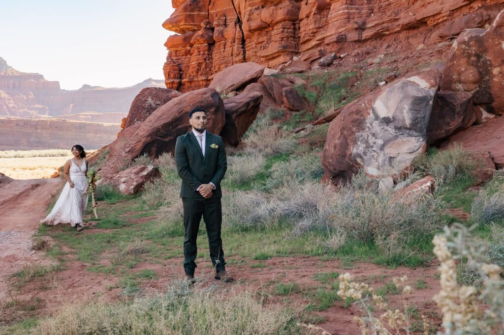 A bride walks behind her groom right before their first look. They are surrounded by red cliffs