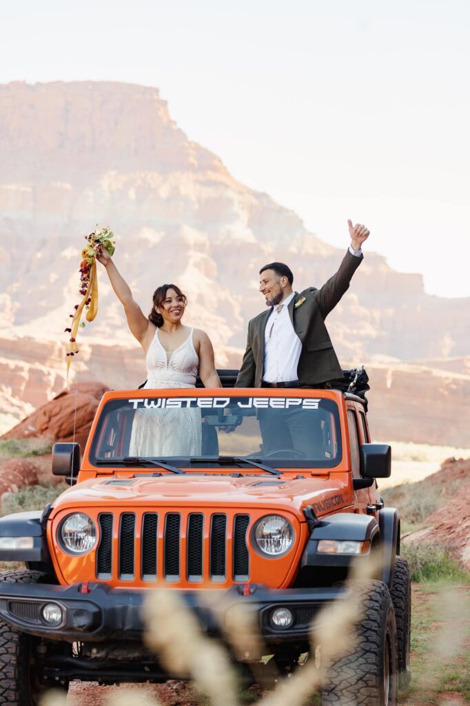 Eloping couple cheering from the top of an orange Jeep in the desert canyon landscape of Moab
