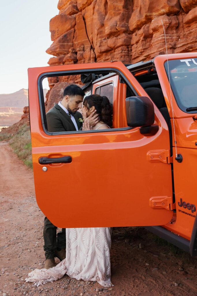 Eloping couple sharing an intimate moment by their orange Jeep along a red rock canyon road in Utah. You can see them through the frame of the Jeep's window.