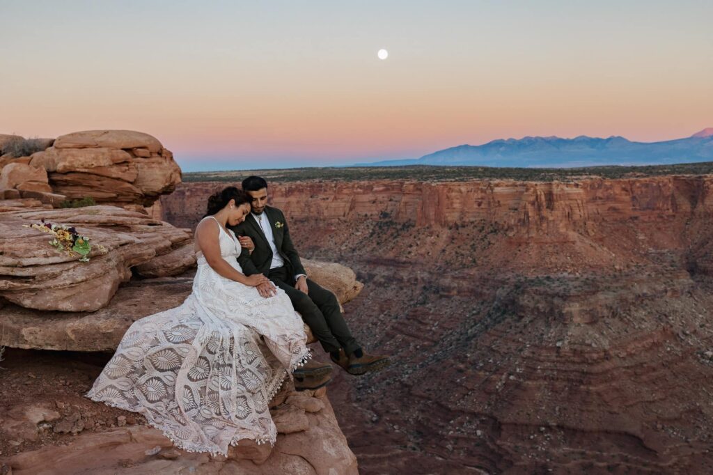 A bride and groom sit on the edge of a canyon holding hands, and taking in the day. The sun is setting and the super moon is rising, and the sky is full of color.