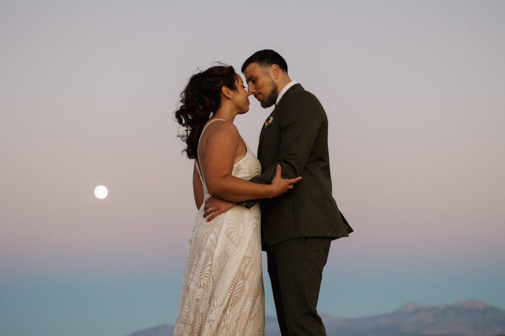 A bride and groom embrace as the sunsets and the super moon rises over the desert cliffs