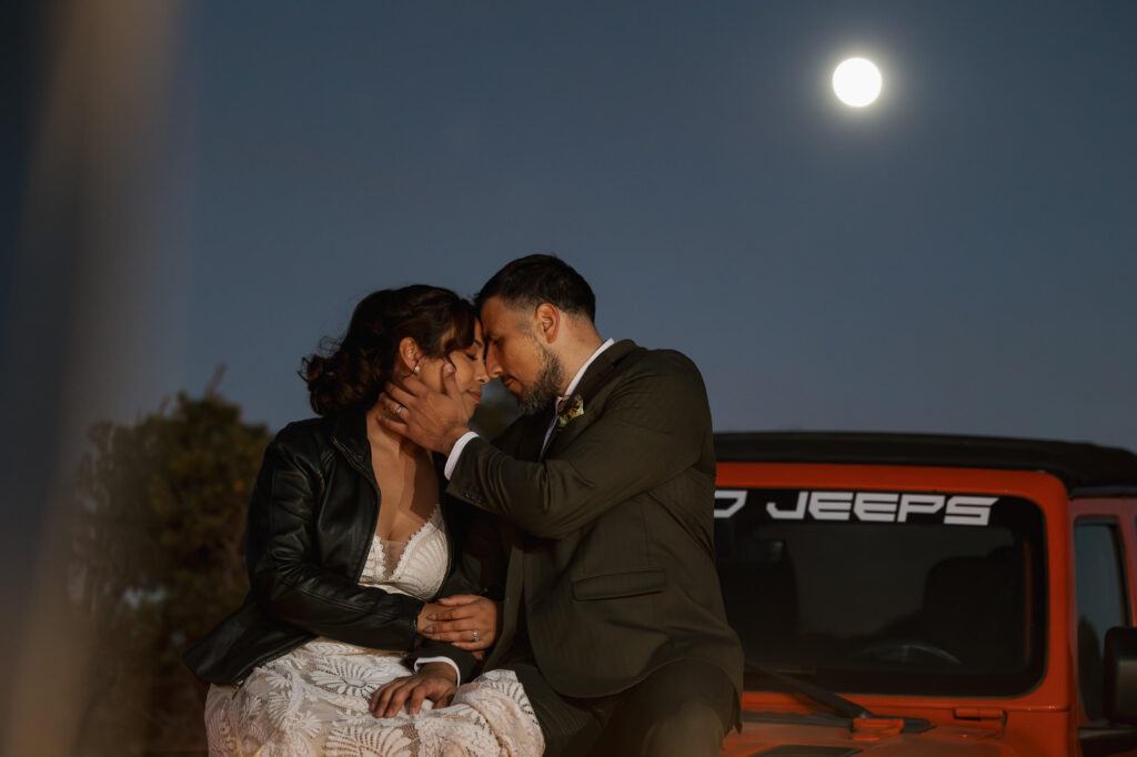 A bride and groom sit on the hood of a jeep with the super moon behind them. The groom gently touches the face of his bride in an intimate moment.