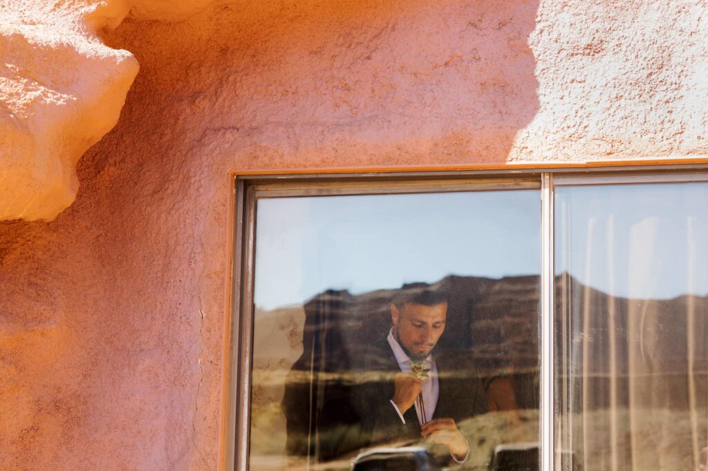 Groom adjusting his boutonniere inside an adobe-style Airbnb with desert cliffs reflected in the window.