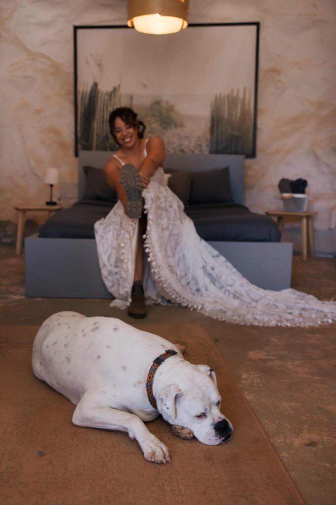 Bride in her wedding dress lacing hiking boots while her white dog rests on the floor in a desert Airbnb.