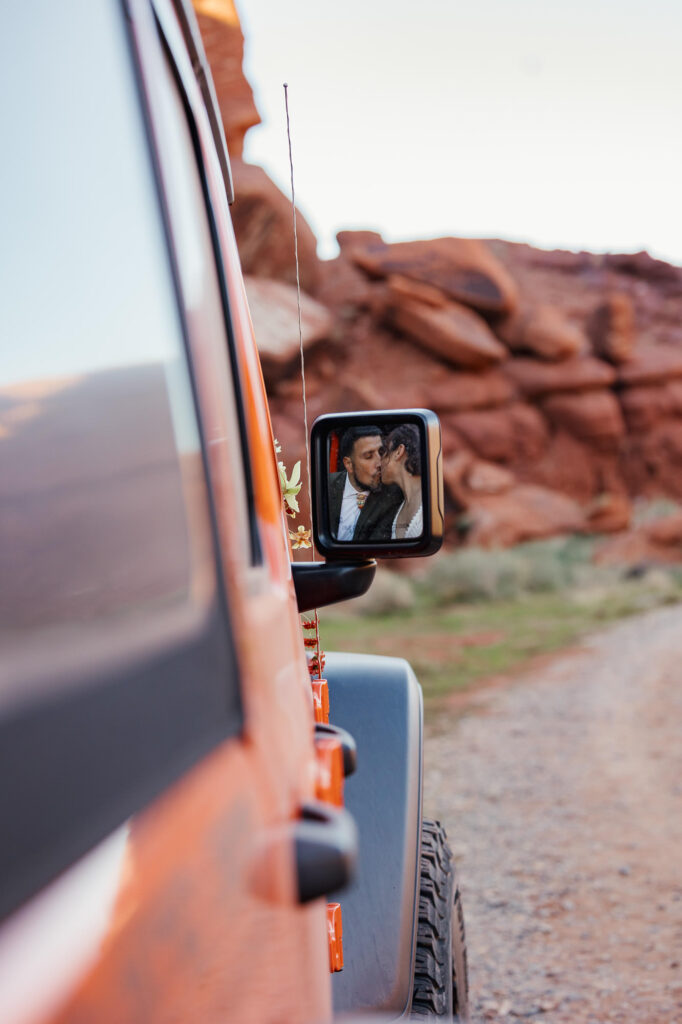 A bride and groom kissing, that you can see in the jeep passenger mirror. There are also red desert cliff in the background.