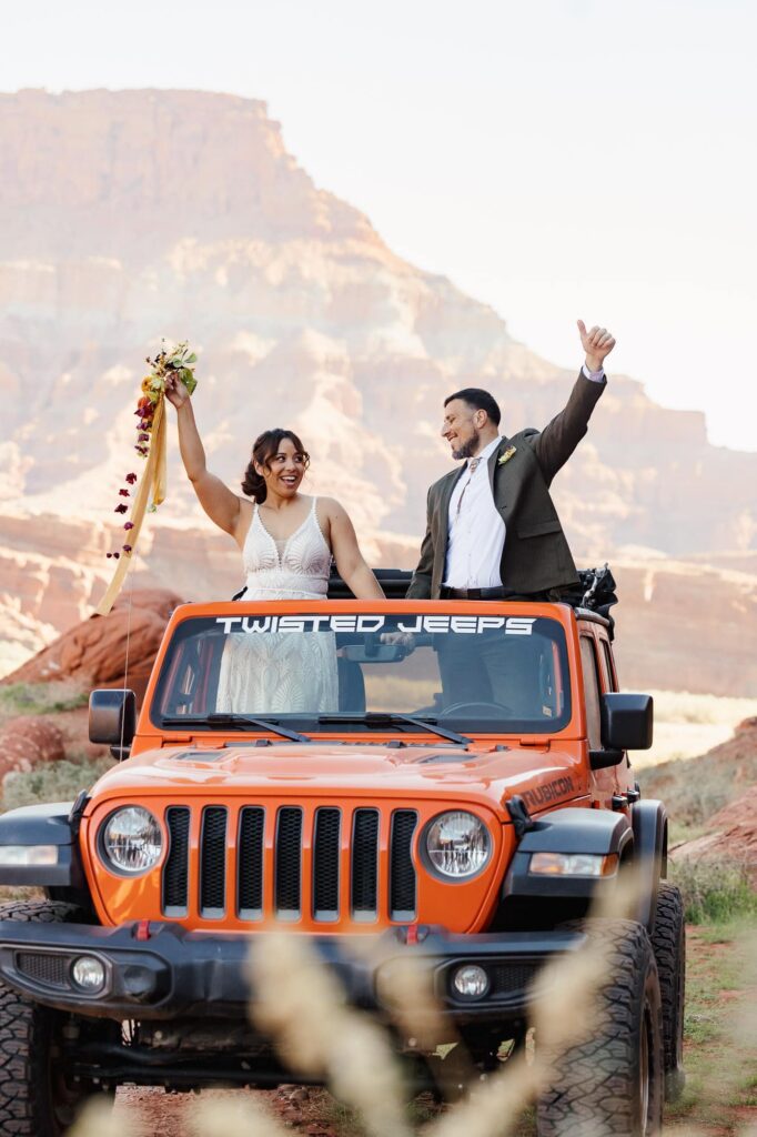 Eloping couple cheering from the top of an orange Jeep in the desert canyon landscape.