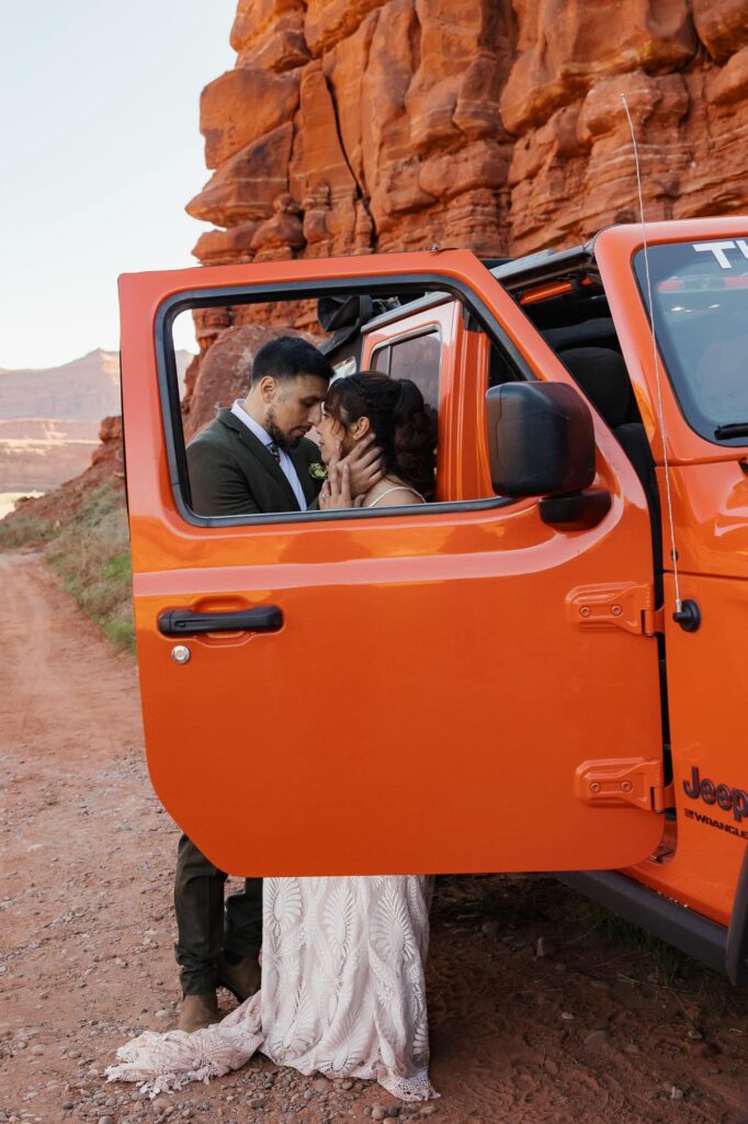 Eloping couple sharing an intimate moment by their orange Jeep along a red rock canyon road in Utah. You can see them through the frame of the Jeep's window.