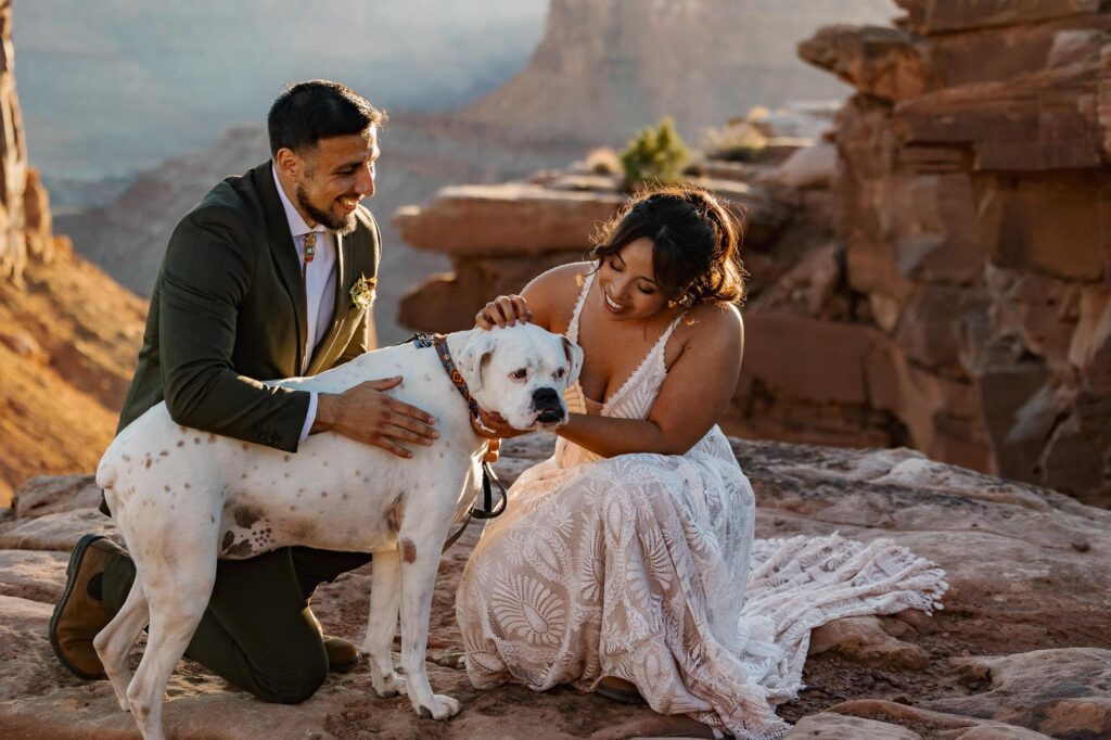 A bride and groom snuggle with their dog at an overlook during their wedding ceremony in Moab