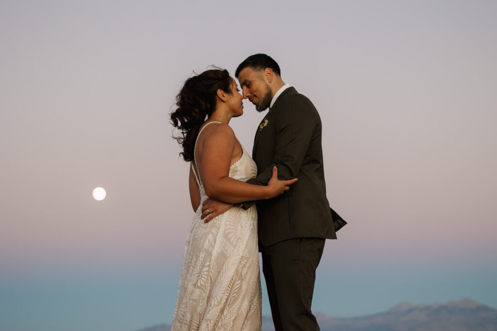 A bride and groom embrace as the sunsets and the super moon rises over the desert cliffs