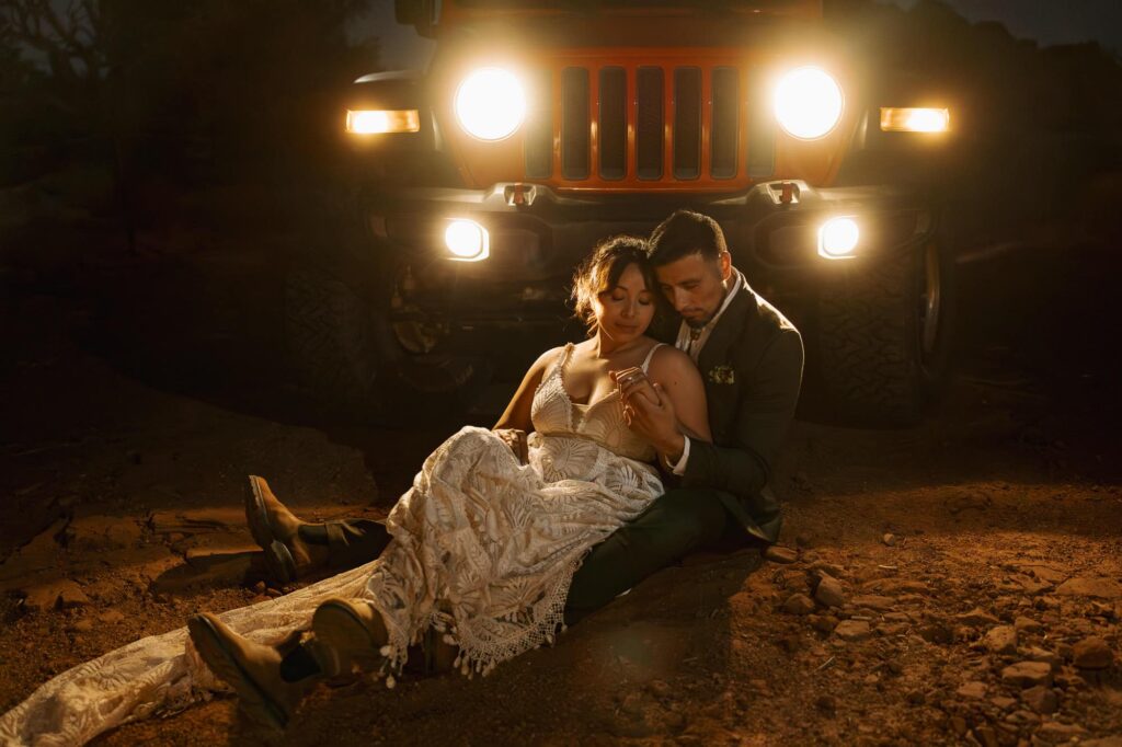 A couple snuggle on the ground lit by the headlights of their jeep as they take in the final moments of their adventure elopement