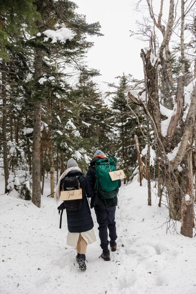 A bride and groom hike back from their ceremony in the snow. They are wearing backpacks with wooden signs that say just eloped on them.