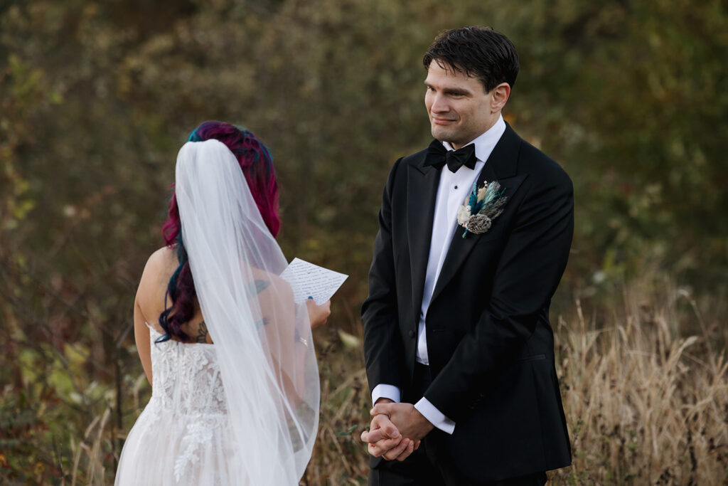 A groom smiles at his bride as she reads her vows to him during their sunrise ceremony at Shenandoah