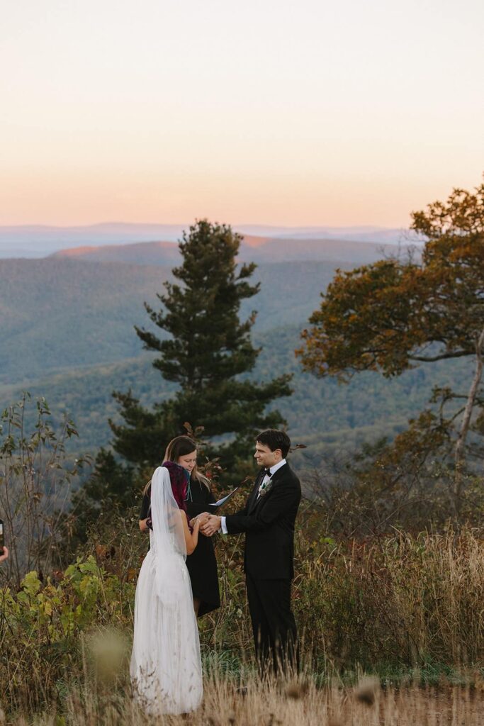 A bride and groom hold hands during their elopement ceremony. You can see the sun rising above the mountains behind them